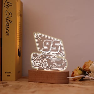 May include: An illuminated LED night light in the shape of a cartoon race car, displaying the number '95' and the name 'Mila'. The light is mounted on a wooden base and emits a warm white glow.