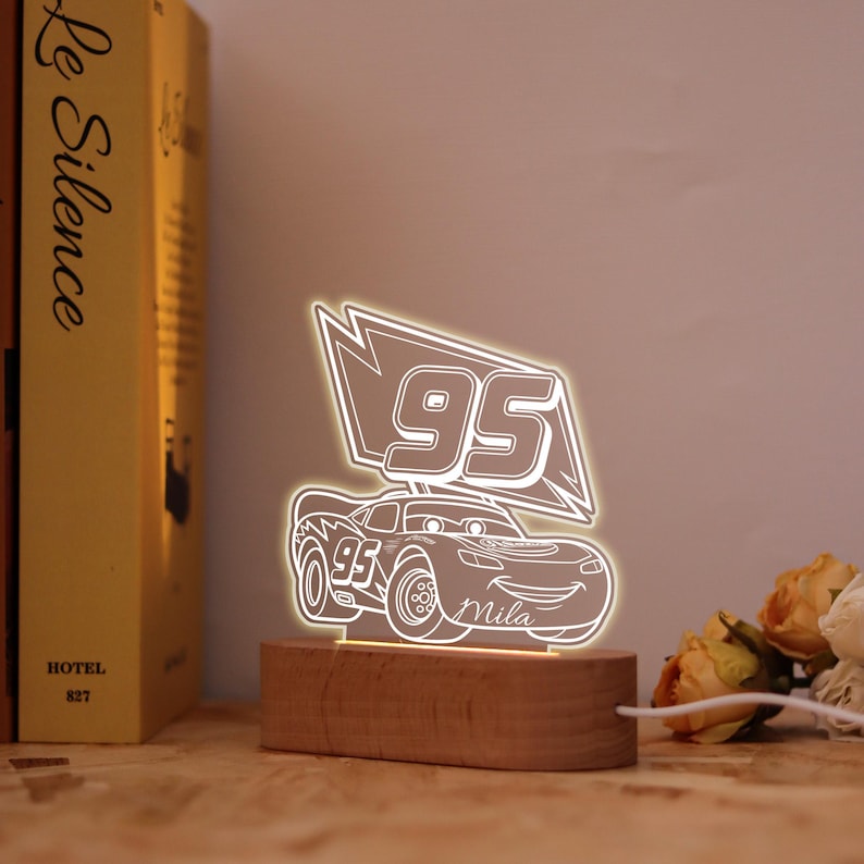 May include: An illuminated LED night light in the shape of a cartoon race car, displaying the number '95' and the name 'Mila'. The light is mounted on a wooden base and emits a warm white glow.