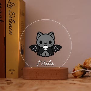 May include: A decorative night light featuring a cartoon bat with a skull on its chest. The bat is gray with black wings and is set against a clear, circular background. The name "Mila" is written below the bat in cursive. The base is made of wood.