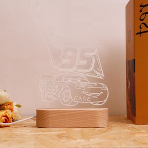 May include: A clear acrylic night light with a race car design, the number 95, and the name "CADE". It rests on a rectangular wooden base. The illuminated design emits a soft glow, perfect for a child's room.