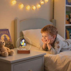 May include: A personalized night light featuring a cartoon character design. The light has a clear acrylic figure with a wooden base. The name "Mila" is printed on the acrylic. The light is on a nightstand next to a bed.