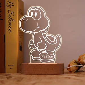 Op de afbeelding: Een oplichtend Yoshi-vormig nachtlampje met de naam "Mila" op de basis. Het licht is gemaakt van helder acryl en staat op een houten voet. Het licht is aangesloten met een wit snoer. Op de achtergrond staan een boek en bloemen.
