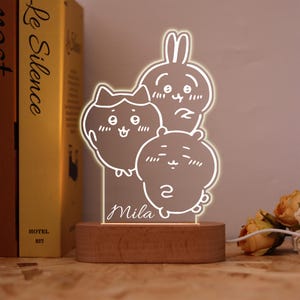 May include: An LED night light with a wooden base, featuring three cartoon characters: a cat, a rabbit, and a bear. The name "Mila" is etched on the front. The light is powered by a cord. The light is approximately 15 cm tall.