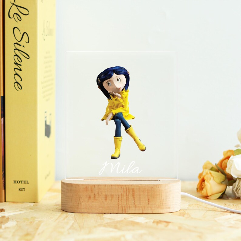 May include: A decorative night light featuring a figurine of a character from the animated film, wearing a yellow raincoat and boots, blue pants, and a dark blue hairstyle. The name "Mila" is written on the light. The light sits on a wooden base.