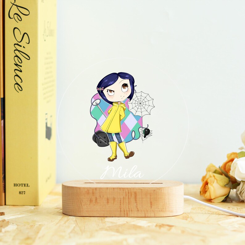 May include: A wooden base supports a clear, circular acrylic night light featuring a cartoon girl in a yellow raincoat. The girl is holding a black bag and a spider web. The name "Mila" is written below the image.