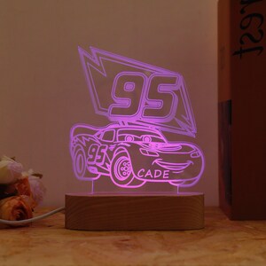 May include: A neon pink LED night light shaped like a cartoon race car with the number 95 and the name CADE. The light sits on a rectangular wooden base and is plugged into an electrical outlet.
