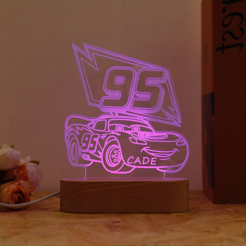 May include: A neon pink LED night light shaped like a cartoon race car with the number 95 and the name CADE. The light sits on a rectangular wooden base and is plugged into an electrical outlet.