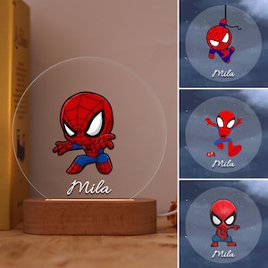 May include: A Spiderman-themed night light with a cartoon Spiderman character in red and blue, and the name "Mila" below. The light sits on a wooden base and has a clear, circular acrylic front.