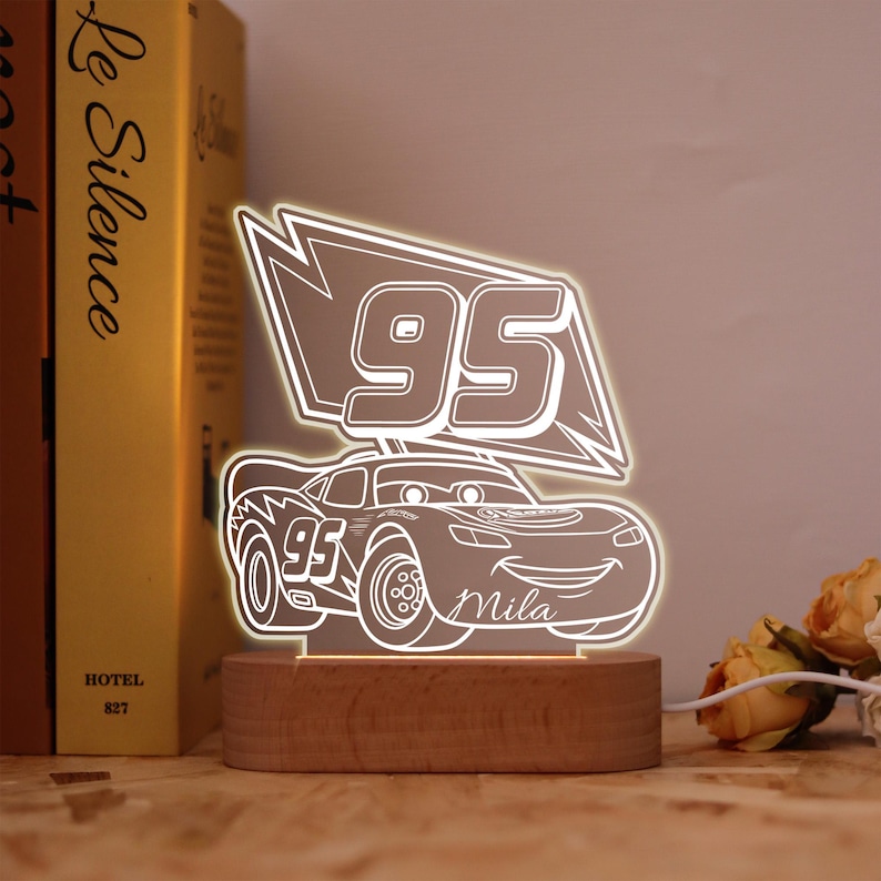 May include: An illuminated LED night light shaped like a cartoon race car. The car displays the number "95" and the name "Mila". The light is mounted on a wooden base and is connected to a power source.