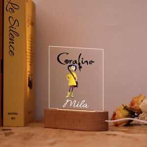 Op de afbeelding: Een LED-nachtlampje met een houten voet, met de namen "Coraline" en "Mila" in script. Het licht schijnt door een helder acryl paneel met een cartoonfiguur in een gele jas. Het licht staat op een houten voet.