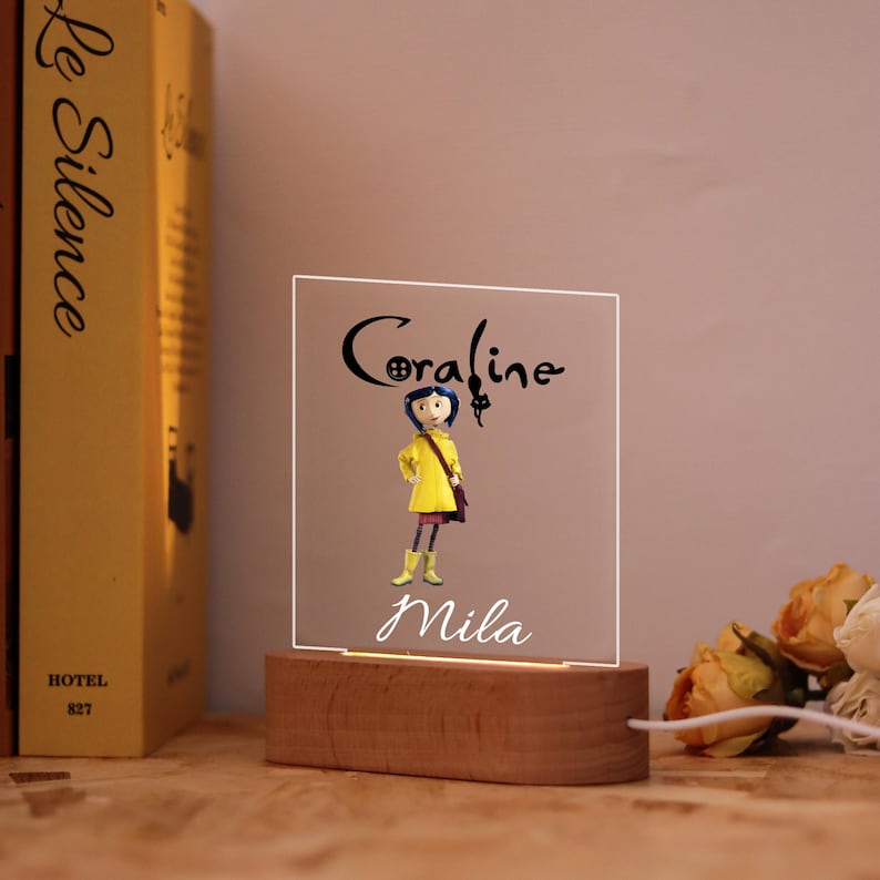 Op de afbeelding: Een LED-nachtlampje met een houten voet, met de namen "Coraline" en "Mila" in script. Het licht schijnt door een helder acryl paneel met een cartoonfiguur in een gele jas. Het licht staat op een houten voet.