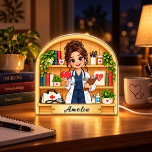 May include: An arched LED night light featuring a cartoon nurse holding a red heart and a clipboard, standing in front of shelves with books, plants, and medical supplies. The base is personalized with the name Amelia in black script.