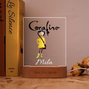 Op de afbeelding: Een decoratief nachtlampje met een stripfiguur in een gele jas, laarzen en een paarse tas. De naam "Coraline" staat boven het personage, met de naam "Mila" eronder. De houten basis luidt "I love you always".