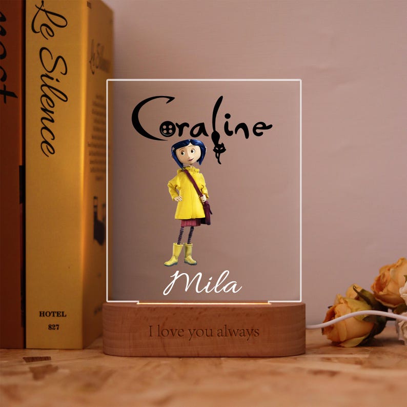 Op de afbeelding: Een decoratief nachtlampje met een stripfiguur in een gele jas, laarzen en een paarse tas. De naam "Coraline" staat boven het personage, met de naam "Mila" eronder. De houten basis luidt "I love you always".