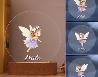 May include: A round, clear acrylic night light featuring a fairy illustration in a purple dress, white wings, and a floral crown. The name "Mila" is written below the fairy. The light sits on a wooden base.