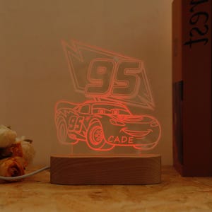 May include: A red LED night light with a race car design, displaying the number "95" and the name "CADE". The light is mounted on a wooden base and is connected to a power source via a cable.