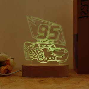 May include: An illuminated LED night light in the shape of a cartoon race car. The design includes the number "95" and the name "CADE". The light sits on a wooden base and emits a soft, warm glow, perfect for a child's bedroom.