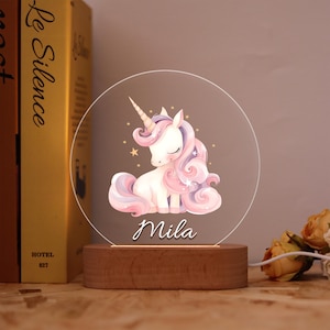 Può includere: Luce notturna decorativa con un design a unicorno. L'unicorno è bianco con criniera e coda rosa e un corno dorato. Il nome "Mila" è scritto sotto. La luce poggia su una base di legno.