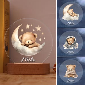 Könnte beinhalten: Nachtlicht mit einem schlafenden Teddybären auf einem Mond und Sternen. Das klare Acryldesign steht auf einem Holzsockel. Der Name "Mila" steht darunter. Zusätzliche Bilder zeigen den Bären auf einer Wolke und in eine Decke gehüllt.