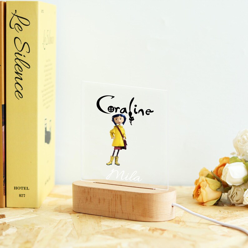 Op de afbeelding: Een nachtlampje met een houten basis en een helder acryl paneel met de naam "Coraline" in script en een illustratie van het personage. De naam "Mila" is ook gegraveerd. Links is een geel boek zichtbaar.