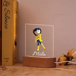 May include: A decorative night light featuring a figurine of a character in a yellow raincoat and blue pants. The figurine is on a clear, illuminated panel with the name "Mila" in script. The base is made of wood, and a book is visible in the background.