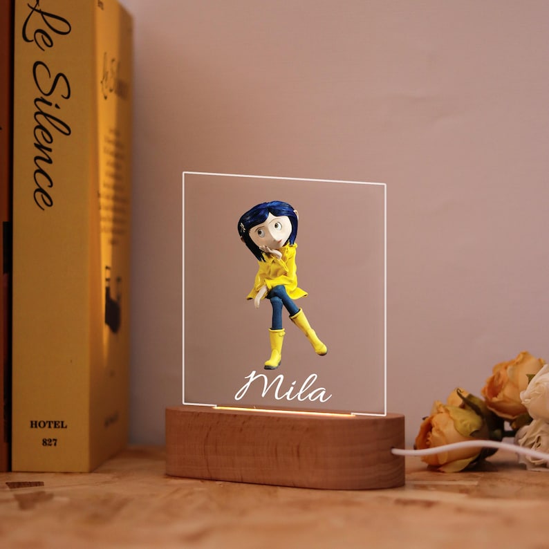 May include: A decorative night light featuring a figurine of a character in a yellow raincoat and blue pants. The figurine is on a clear, illuminated panel with the name "Mila" in script. The base is made of wood, and a book is visible in the background.
