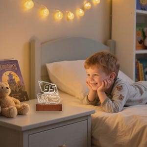 May include: A lit-up night light with a car design and the number "95" on a wooden base. The light is on a bedside table, with a child looking at the light.
