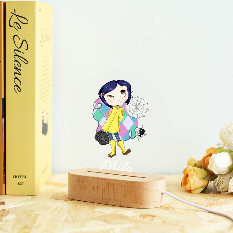May include: A clear acrylic night light featuring a cartoon illustration of a girl in a yellow raincoat, holding a black bag and standing near a spider web. The light sits on a wooden base with the name "Mila" and a power cord.