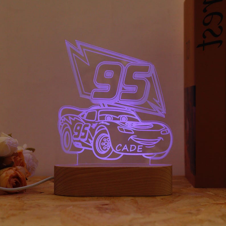 May include: A purple LED night light with a cartoon race car design. The car features the number "95" and the name "CADE". The light is mounted on a rectangular wooden base and is plugged in.