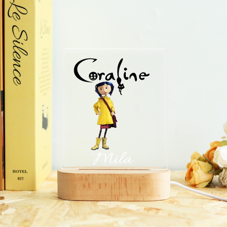 Op de afbeelding: Decoratief nachtlampje met het personage Coraline uit de animatiefilm. Het ontwerp bevat de naam "Coraline" in een gestileerd lettertype, de karakterillustratie en de naam "Mila". Het licht staat op een houten voet.