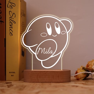 May include: A glowing white outline of a cartoon character, with the name "Mila" in script, on a wooden base. The background includes a book and flowers. The light emits a soft glow.
