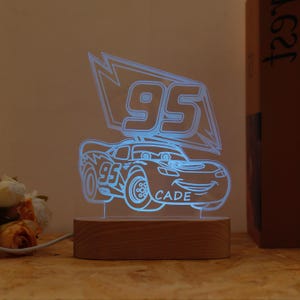 May include: A blue LED night light shaped like a cartoon race car with the number 95 and the name "CADE". The light sits on a rectangular wooden base and is plugged in with a white cord. The light is a fun decorative item.