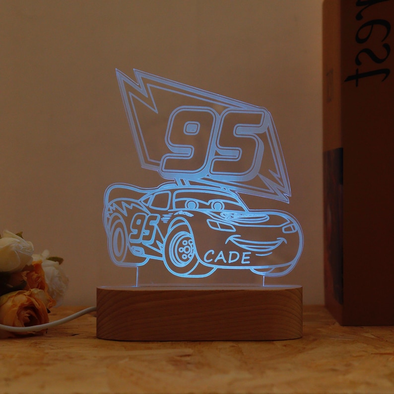 May include: A blue LED night light shaped like a cartoon race car with the number 95 and the name "CADE". The light sits on a rectangular wooden base and is plugged in with a white cord. The light is a fun decorative item.