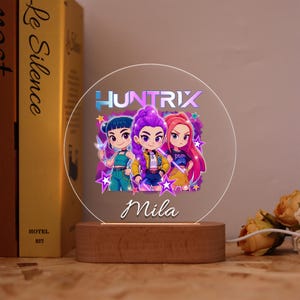 May include: A round, illuminated night light with a wooden base. The light features a cartoon image of three girls with the text "HUNTRIX" and the name "Mila". The girls have blue, purple, and red hair. The light is powered by a white cord.
