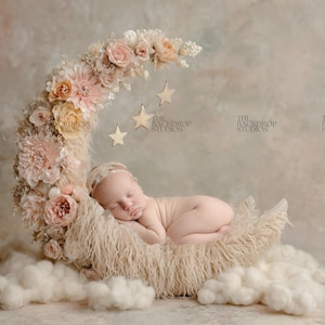 May include: A newborn baby sleeps peacefully on a fluffy, cream-colored moon prop, adorned with flowers and hanging stars. The backdrop is a soft, neutral color, creating a serene and dreamy scene for newborn photography.