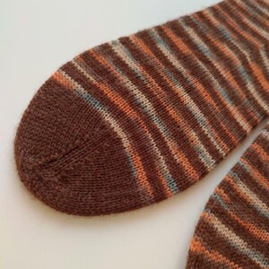 May include: A pair of knitted socks featuring a striped pattern in shades of dark brown, burnt orange, and light blue. The fabric has a soft, fuzzy texture with a rounded toe construction and a consistent knit stitch throughout the design.