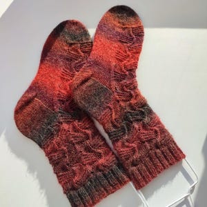 May include: A pair of hand-knitted socks featuring a variegated colour palette of deep red, orange, and dark charcoal grey. The socks display a textured cable knit pattern throughout the leg and foot, finished with a ribbed cuff at the top opening.