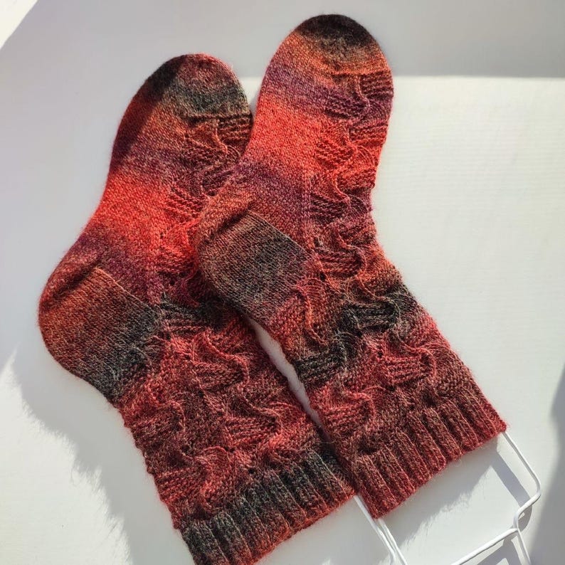 May include: A pair of hand-knitted socks featuring a variegated colour palette of deep red, orange, and dark charcoal grey. The socks display a textured cable knit pattern throughout the leg and foot, finished with a ribbed cuff at the top opening.