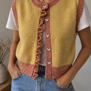 May include: A mustard yellow knit vest featuring a pink ribbed trim along the neckline, armholes, and hem. The front closure includes a ruffled pink edge and five silver butterfly-shaped buttons over a white t-shirt and blue denim jeans.
