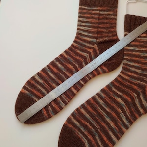 May include: A pair of hand-knitted socks featuring horizontal stripes in shades of brown, orange, and light blue. A metal ruler is placed diagonally across one sock, showing a length measurement of approximately 29 cm from toe to heel.
