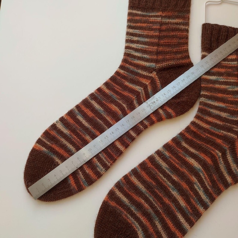 May include: A pair of hand-knitted socks featuring horizontal stripes in shades of brown, orange, and light blue. A metal ruler is placed diagonally across one sock, showing a length measurement of approximately 29 cm from toe to heel.