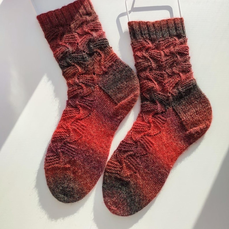May include: A pair of hand-knitted socks featuring a variegated colour palette of deep red, orange, and dark charcoal grey. The socks display a textured, wavy cable knit pattern throughout the leg and foot, finished with a ribbed cuff at the top.