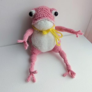 May include: A knitted frog toy in pink and white, with a yellow scarf and large black eyes. The frog has long, articulated limbs. The body is white, while the head and limbs are pink. The toy is handmade and sits on a white surface.