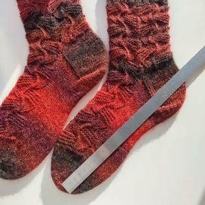 May include: A pair of hand-knitted socks featuring a variegated colour pattern of red, orange, and dark brown tones. The socks have a textured, raised stitch design and are positioned next to a metal ruler showing a length of 23 cm.