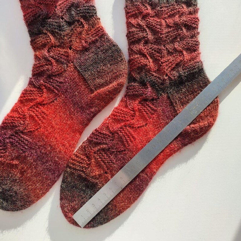 May include: A pair of hand-knitted socks featuring a variegated colour pattern of red, orange, and dark brown tones. The socks have a textured, raised stitch design and are positioned next to a metal ruler showing a length of 23 cm.
