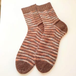 May include: A pair of knitted socks featuring a ribbed cuff and a variegated striped pattern in shades of brown, peach, and light blue. The socks have a solid brown heel and toe section, with a consistent knit texture throughout the entire garment.