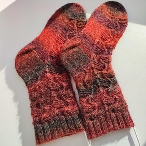 May include: A pair of hand-knitted socks featuring a variegated colour palette of deep red, orange, and dark charcoal grey. The socks display a textured, wavy cable knit pattern throughout the leg and foot, finished with a ribbed cuff at the top.