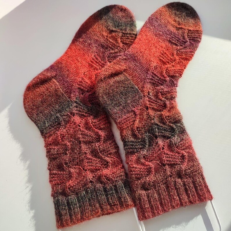 May include: A pair of hand-knitted socks featuring a variegated colour palette of deep red, orange, and dark charcoal grey. The socks display a textured, wavy cable knit pattern throughout the leg and foot, finished with a ribbed cuff at the top.