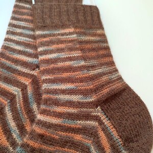 May include: A pair of hand-knitted socks featuring a variegated yarn pattern in shades of brown, soft orange, and light blue. The socks have a ribbed cuff at the top and a solid brown heel and toe section, showcasing a consistent knit texture throughout.