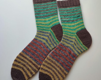 May include: A pair of knit socks featuring horizontal stripes in shades of brown, green, yellow, and orange. The socks have solid brown cuffs, heels, and toes, and are displayed on metal wire hangers against a plain, light-colored background.